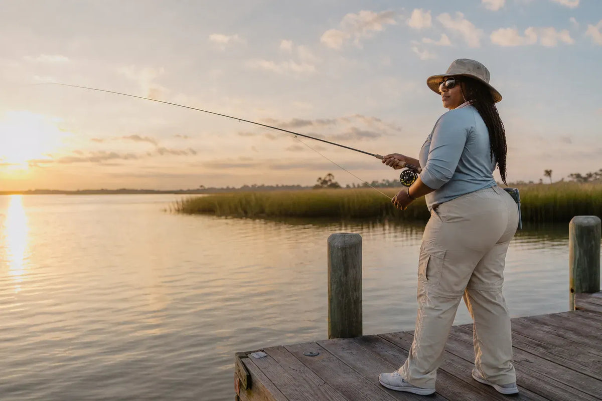 A woman fly fishing from a weathered dock at golden hour, with calm marsh water behind her